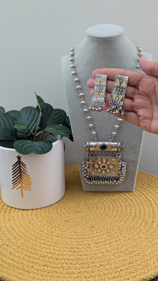 Oxidized silver long necklace with a statement pendant displayed on a mannequin, accompanied by matching earrings, against a white background with a plant to the side.
