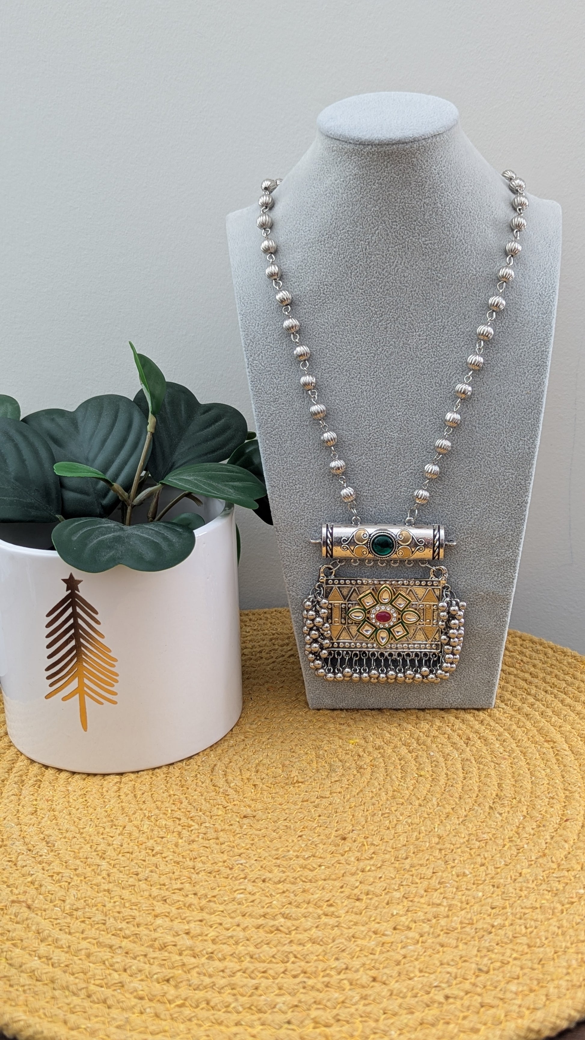 Oxidized silver long necklace with a statement pendant displayed on a mannequin, accompanied by matching earrings, against a white background with a plant to the side.