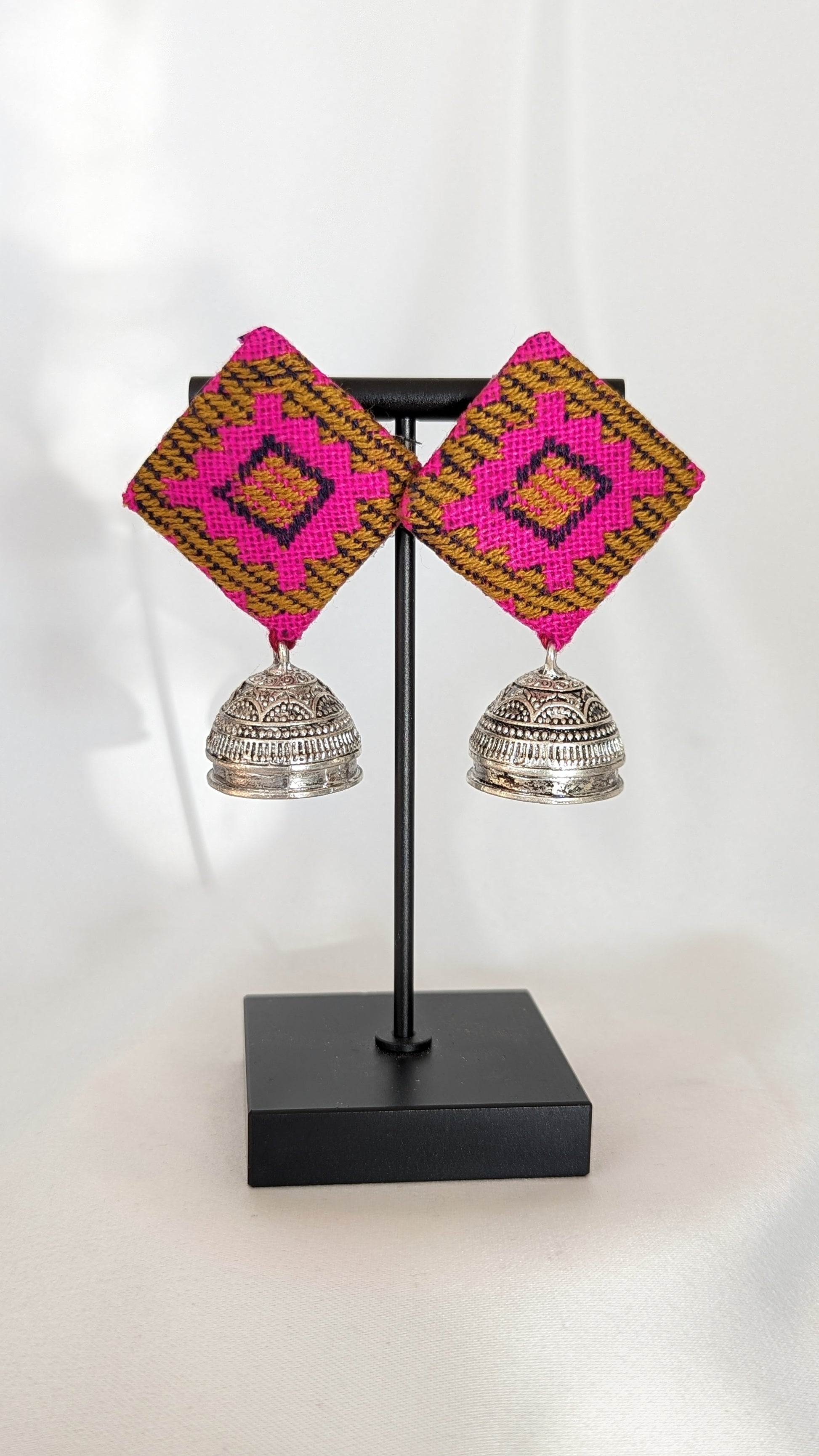 A pair of pink ikat fabric earrings displayed on a stand, with the earrings featuring a geometric pattern and silver-colored bell-like attachments. Pick-up in Ajax Ontario. Worldwide shipping available. Perfect for wedding, festival jewelry, indian Jewelry, kids jewelry, kundan jewelry, gifting, costume jewelry, fashion jewelry, Eid, onam, Diwali, karwachauth, navratri. Free shipping available. Jewelry for women. Indian jewelry in ajax durham region.