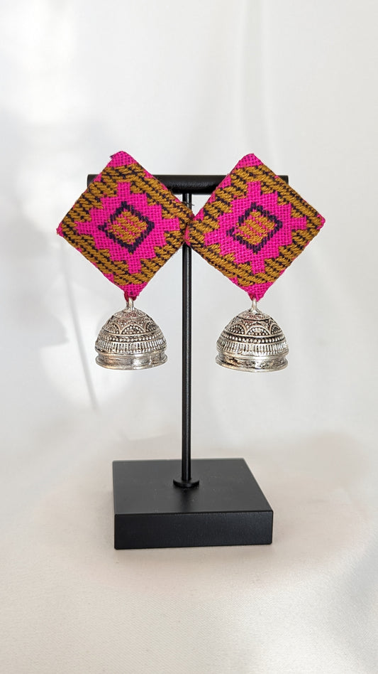 A pair of pink ikat fabric earrings displayed on a stand, with the earrings featuring a geometric pattern and silver-colored bell-like attachments. Pick-up in Ajax Ontario. Worldwide shipping available. Perfect for wedding, festival jewelry, indian Jewelry, kids jewelry, kundan jewelry, gifting, costume jewelry, fashion jewelry, Eid, onam, Diwali, karwachauth, navratri. Free shipping available. Jewelry for women. Indian jewelry in ajax durham region.